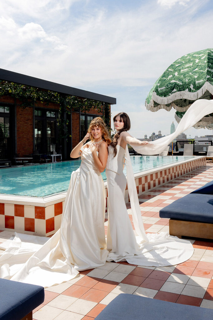 Two brides in flowing white wedding gowns pose poolside on a rooftop terrace, with one lifting a sheer veil as it drapes in the breeze.
