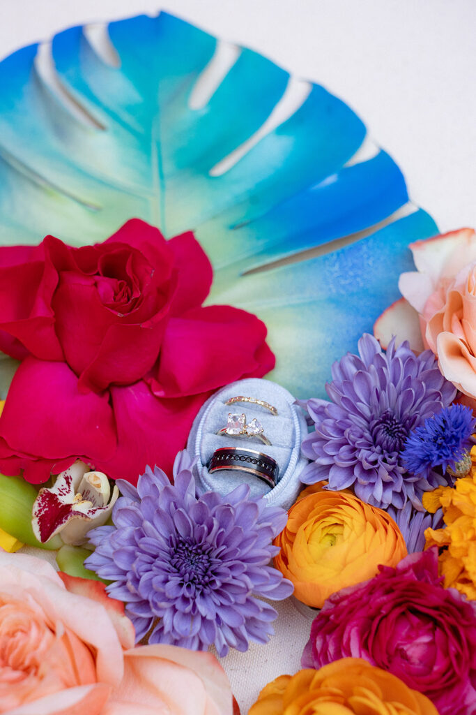 Wedding rings displayed in a blue velvet ring box surrounded by vibrant, colorful flowers and tropical leaf accents.