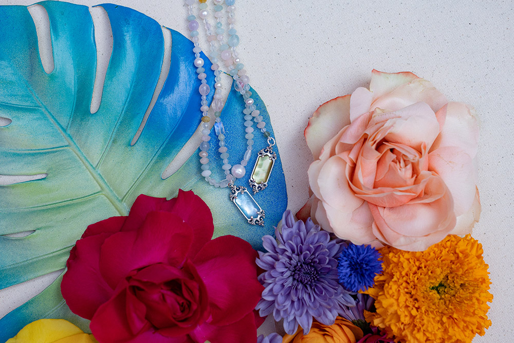 Beaded necklaces with gemstone pendants styled on a blue leaf alongside colorful flowers on a neutral fabric background.