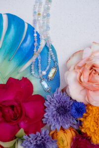 Beaded necklaces with gemstone pendants styled on a blue leaf alongside colorful flowers on a neutral fabric background.