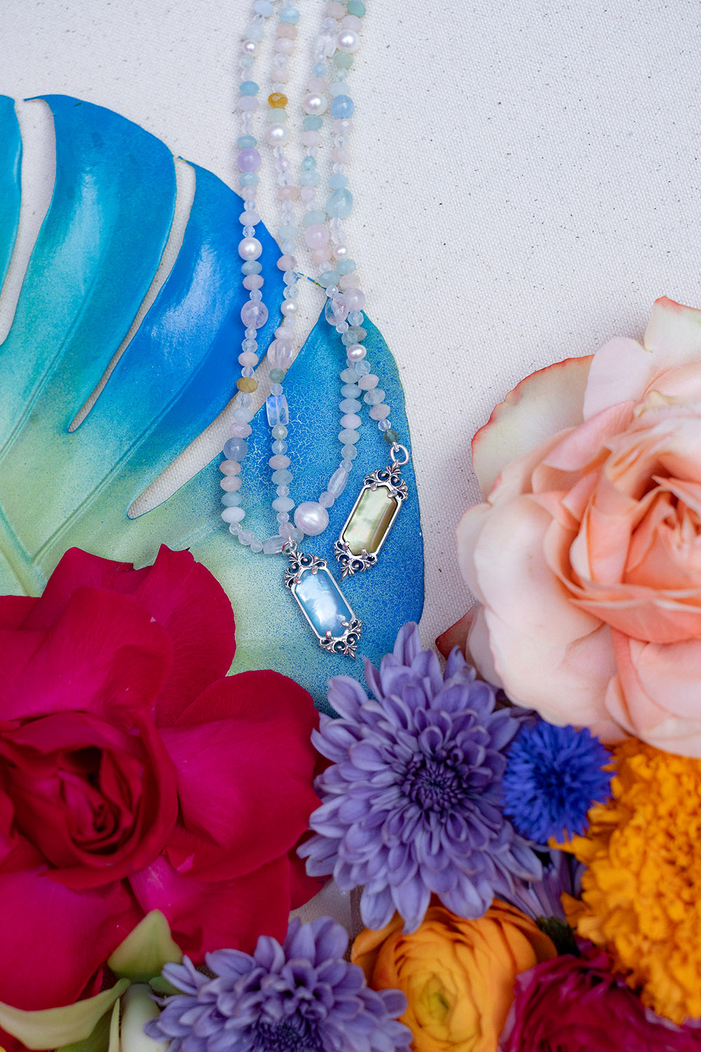 Beaded necklaces with gemstone pendants styled on a blue leaf alongside colorful flowers on a neutral fabric background.