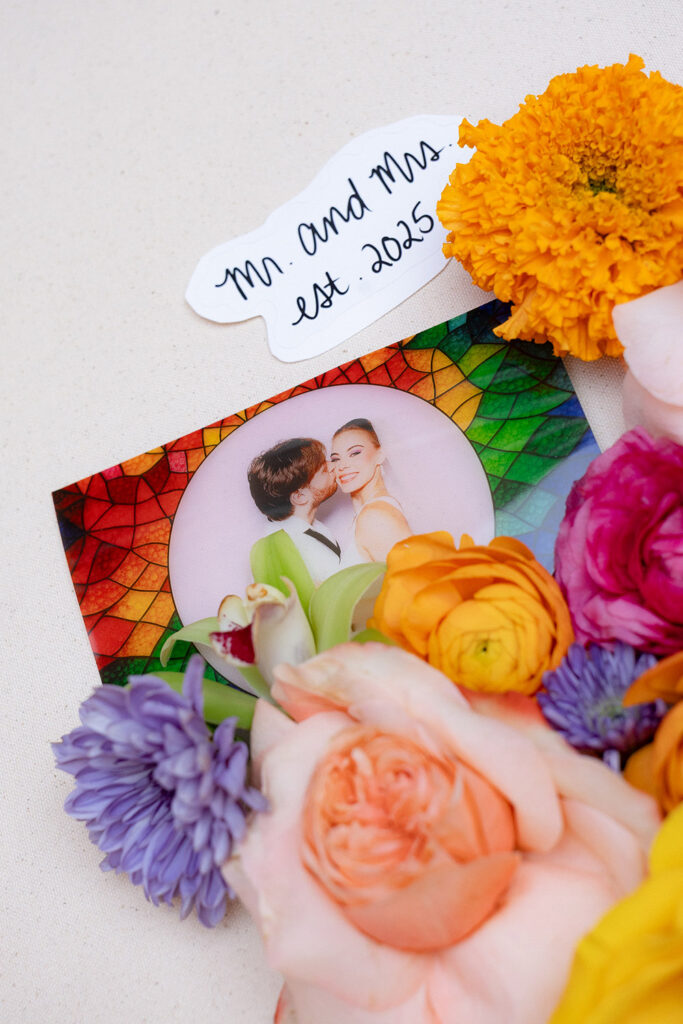 Close-up of colorful wedding florals surrounding a custom card reading “Mr. and Mrs., est. 2025” with a circular photo of the couple.
