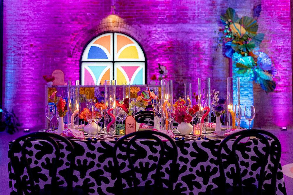 Vibrant wedding reception table with black-and-white patterned linens, colorful florals, tall glass candles, and a stained-glass–style window backdrop under pink and purple uplighting.