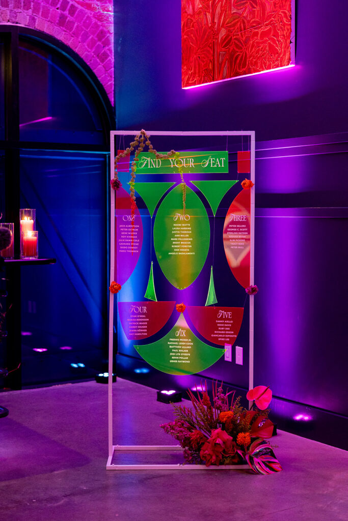 Colorful acrylic escort card display with modern geometric design, guest names, and floral accents, illuminated by purple and pink uplighting against exposed brick walls.