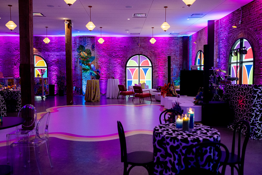Colorful indoor wedding reception space with purple and pink uplighting, arched windows, lounge seating, and a central dance floor.