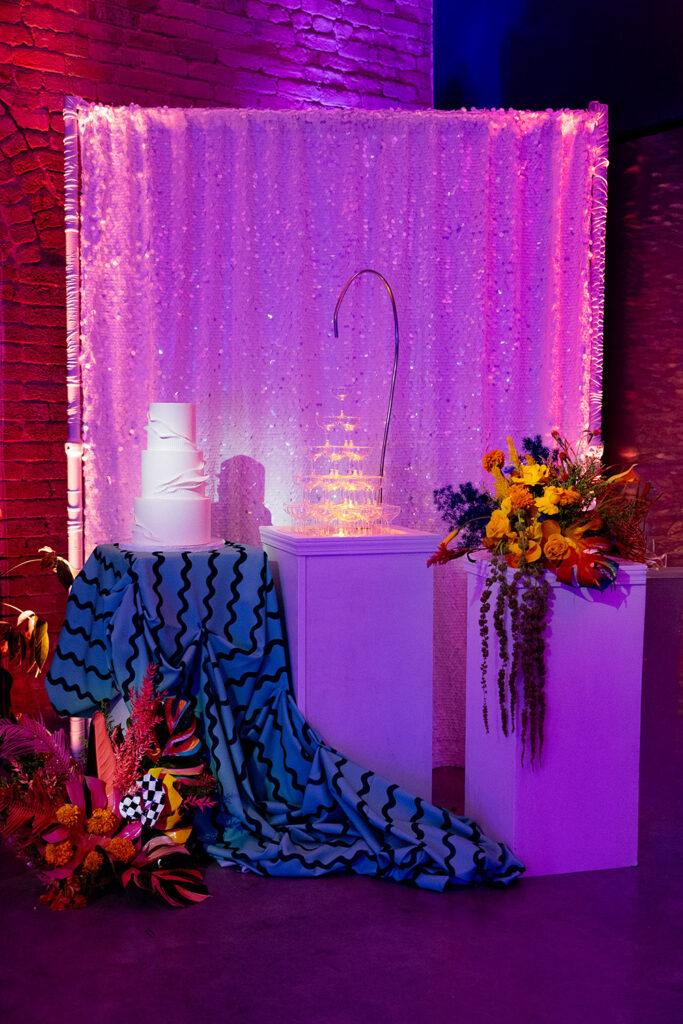 Modern wedding cake display with a white sculptural cake, champagne tower, bold floral arrangement, and shimmering sequin backdrop lit in pink and purple tones.