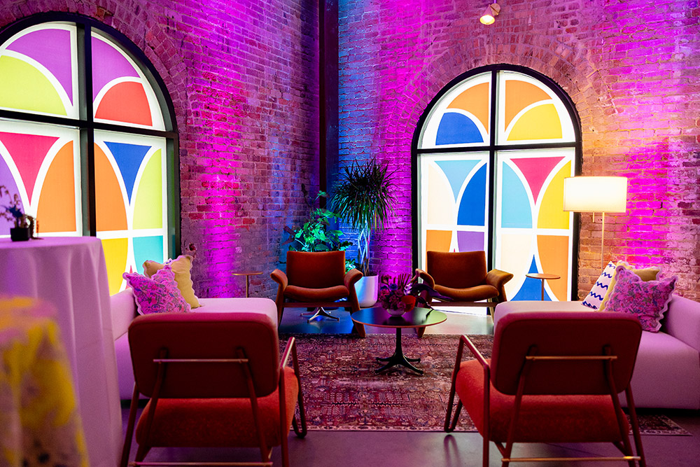 Lounge seating vignette with colorful upholstered chairs, patterned pillows, stained-glass style arched windows, and pink and purple uplighting against exposed brick walls.