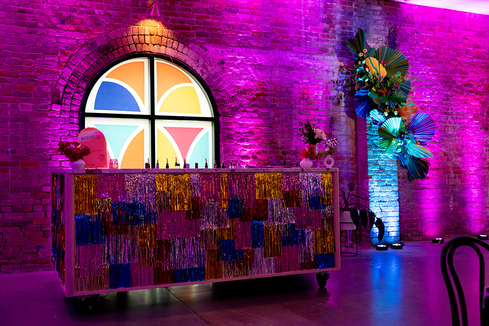 Colorful wedding bar setup with shimmering fringe panels, purple uplighting, brick walls, and a stained-glass style arched window backdrop.
