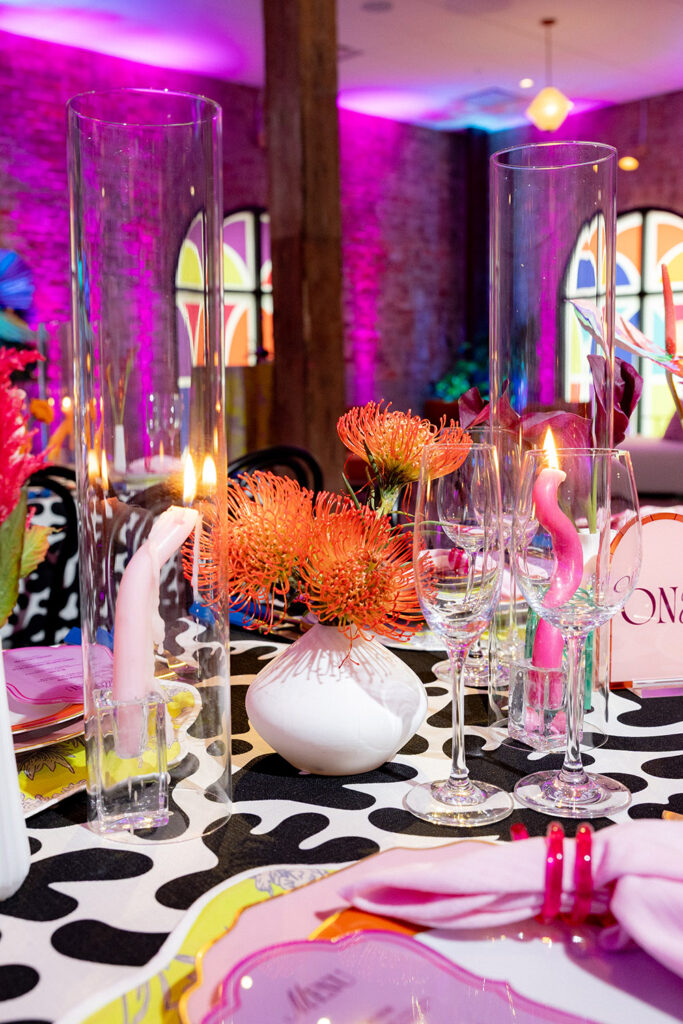 Vibrant reception tablescape with glass cylinder candles, colorful floral centerpieces, sculptural candles, and modern place settings under pink and purple uplighting.