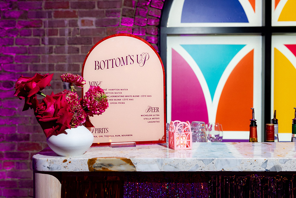 Colorful wedding bar setup with a “Bottom’s Up” drink menu, terrazzo bar top, red florals, and stained-glass–style window backdrop with purple lighting.