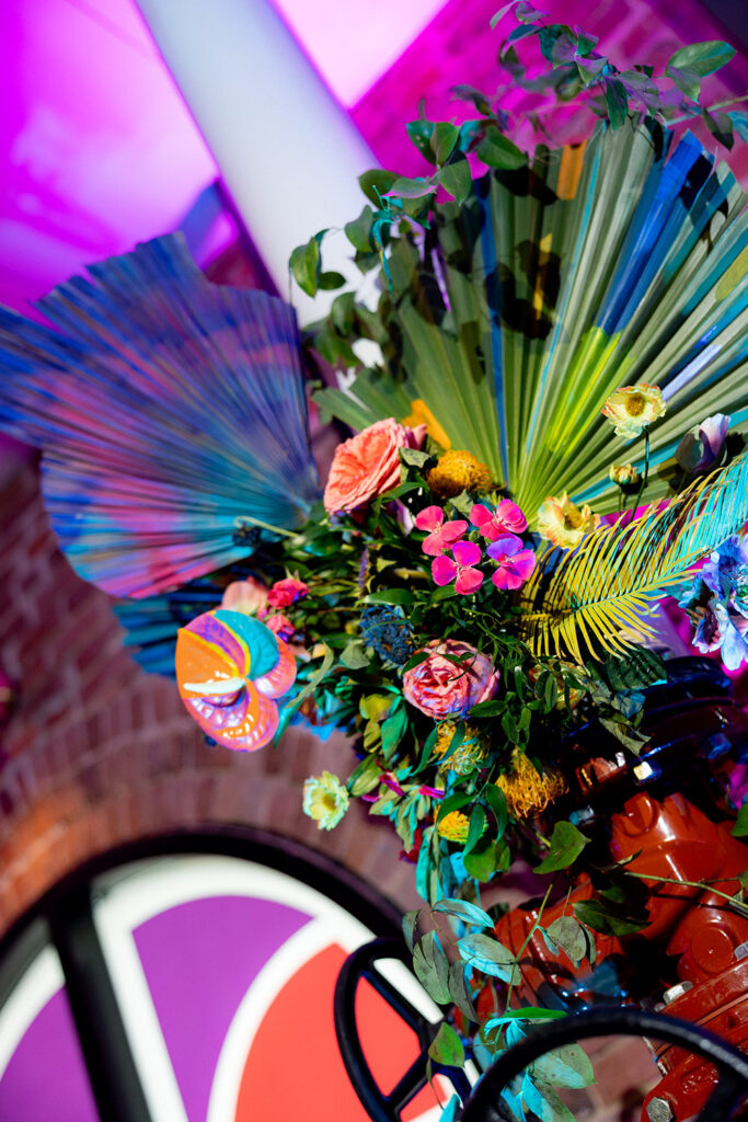 Vibrant floral arrangement with colorful fan palms, tropical blooms, and greenery displayed against exposed brick with pink and purple lighting.