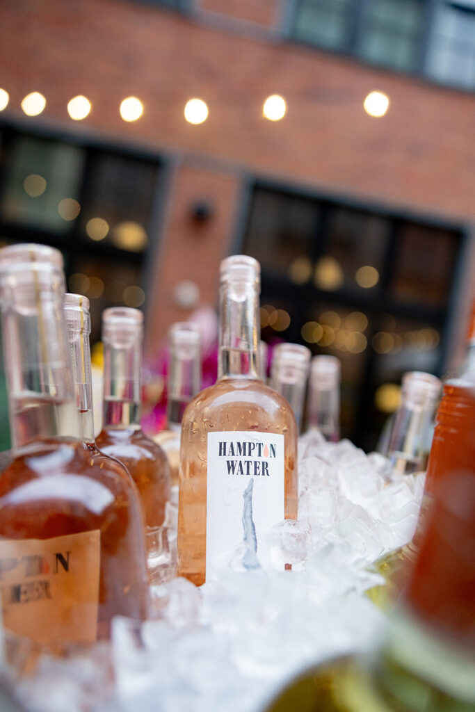 Bottles of Hampton Water rosé chilled on ice at an outdoor wedding bar, with café lights and brick building in the background.