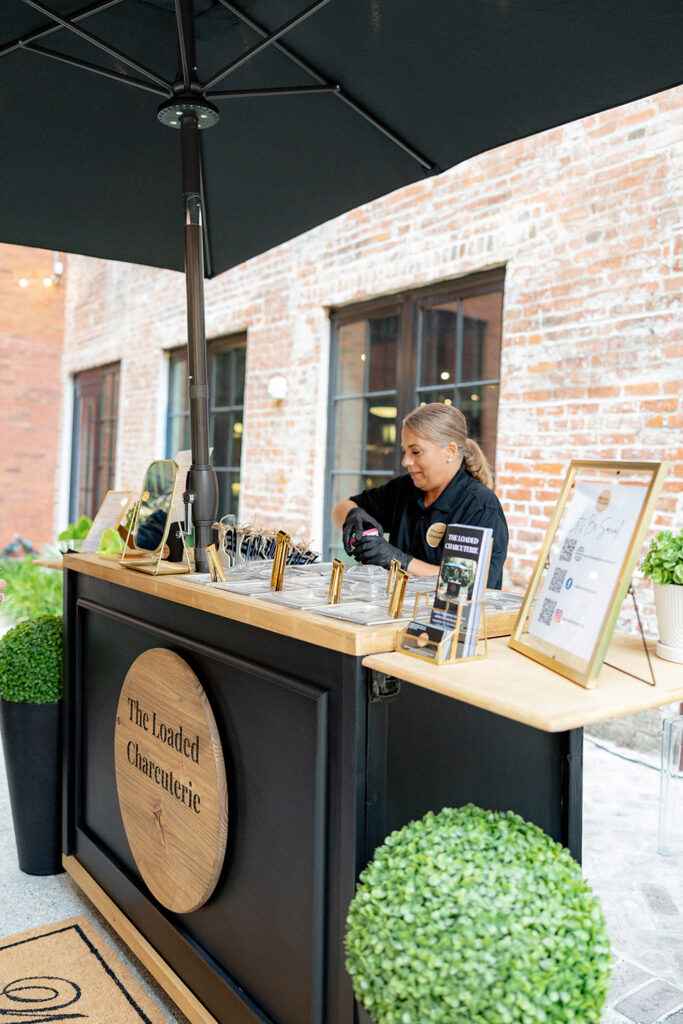 Wedding charcuterie station with a black cart and umbrella, staffed by a vendor preparing individual servings in an outdoor brick courtyard.