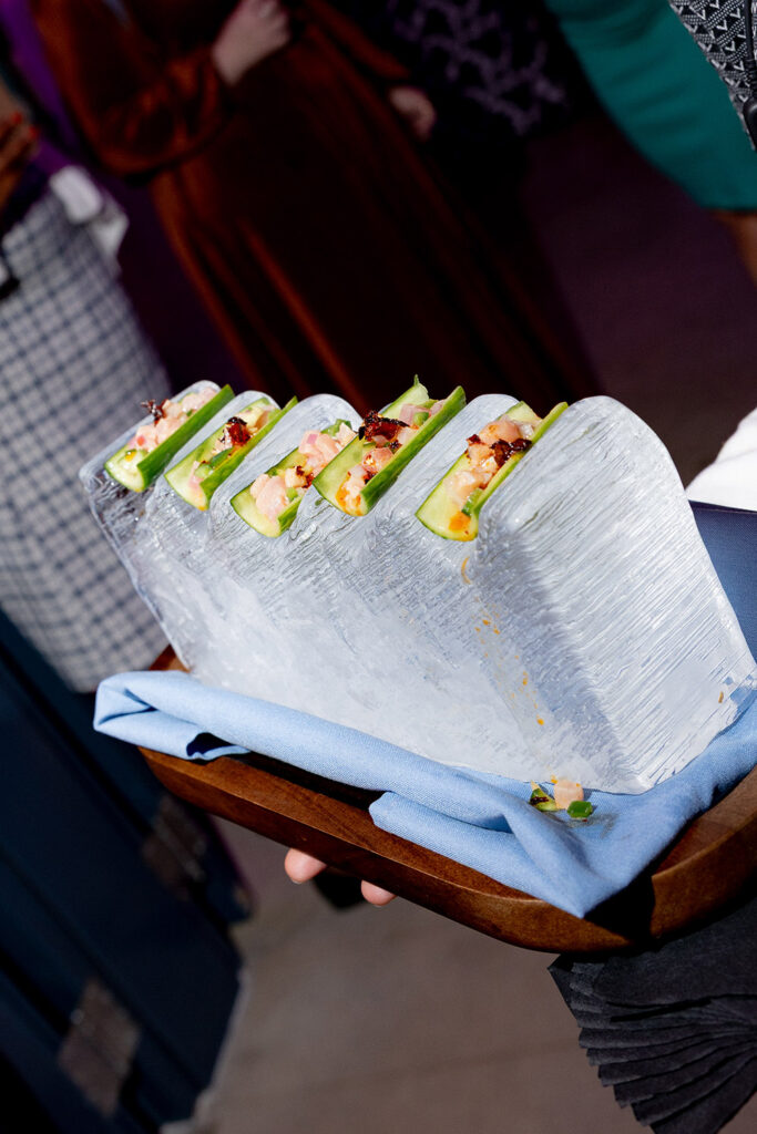 Cucumber bites filled with seafood, displayed in an ice block and served on a wooden tray at a wedding reception.
