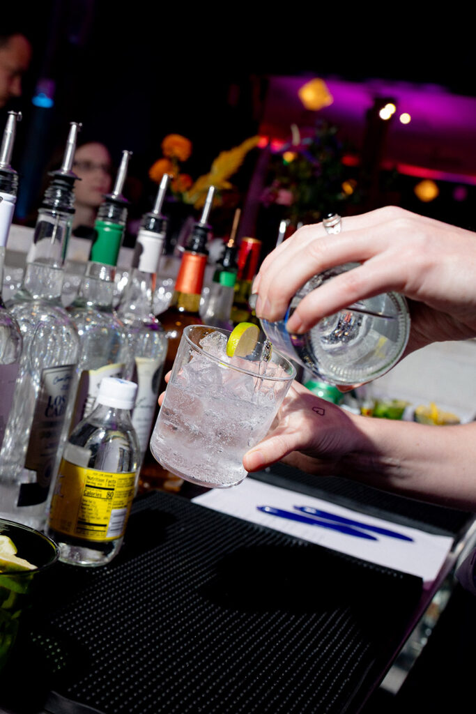 Bartender pouring a clear cocktail over ice with a lime garnish at a wedding reception bar.