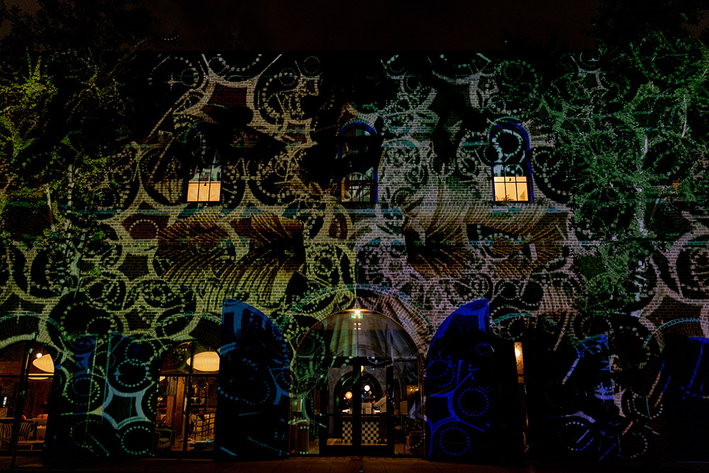 Nighttime exterior of a brick venue illuminated with colorful patterned light projections across the façade and arched windows.