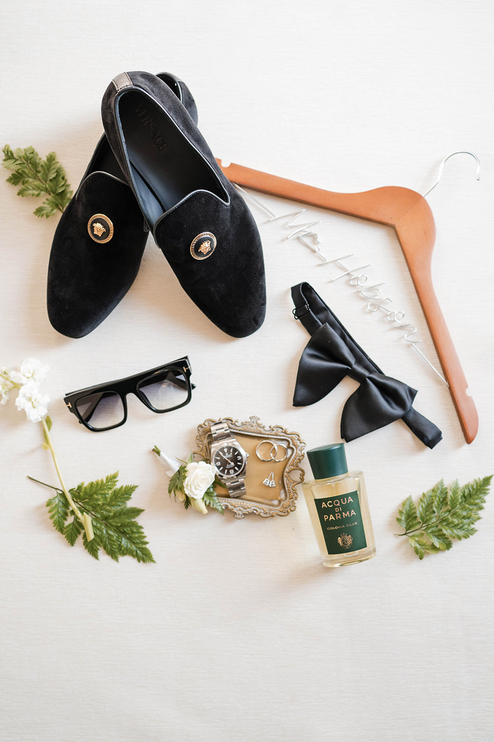 Flat lay of groom accessories including black loafers, bow tie, sunglasses, wristwatch, wedding rings, cologne, and suit hanger on a light background.