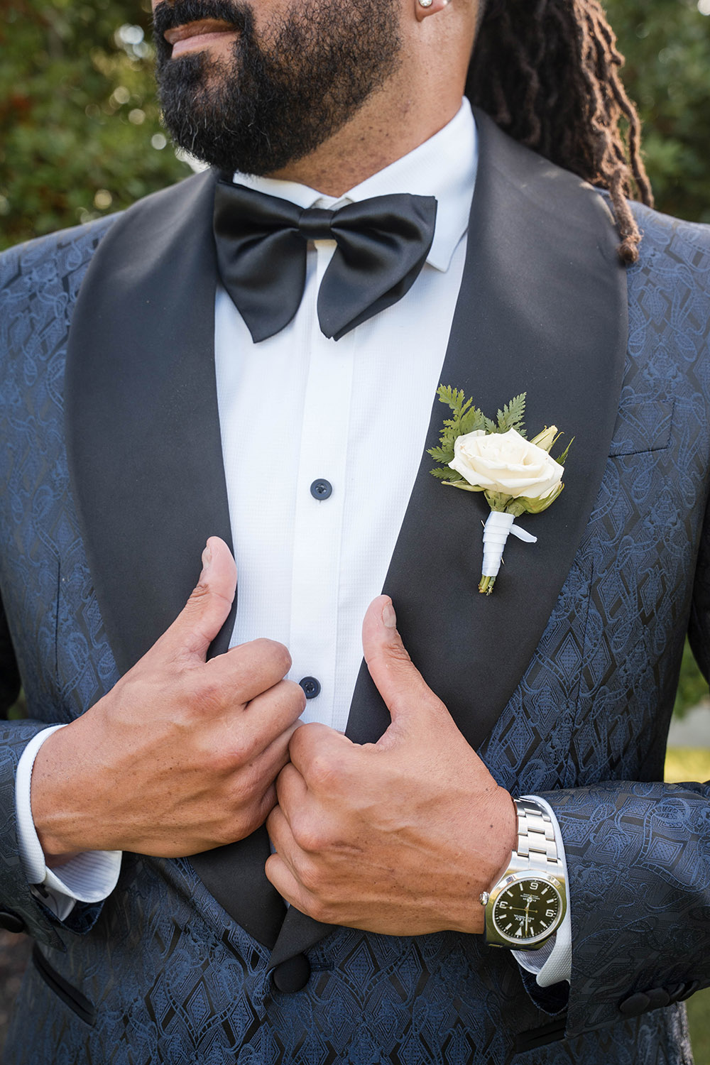 Close-up of the groom adjusting his suit jacket, wearing a black bow tie, white boutonniere, and wristwatch.