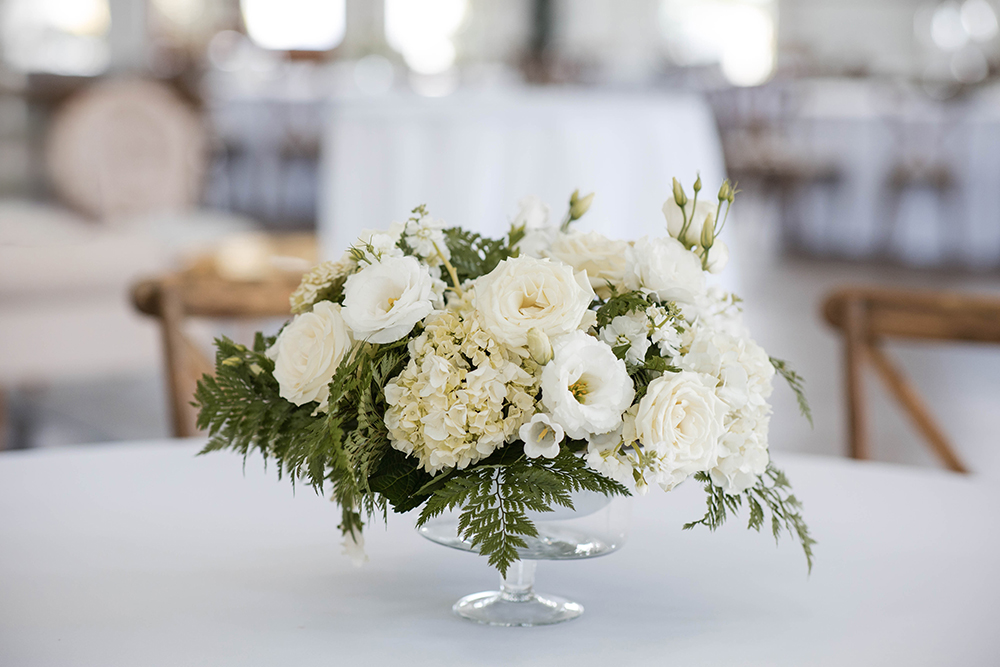 Low floral centerpiece of white roses and greenery arranged in a glass bowl on a round reception table.