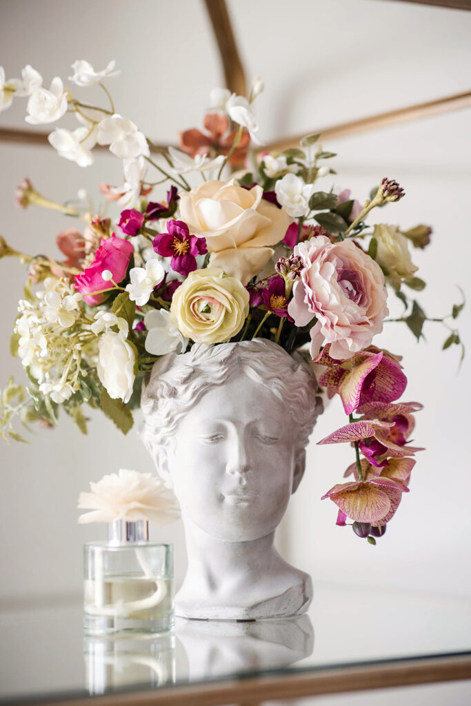 Decorative floral arrangement displayed in a white sculpted bust, featuring pastel roses, orchids, and greenery on a tabletop.