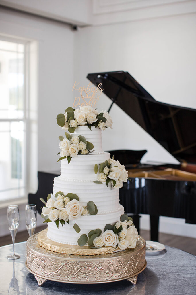 Three-tier white wedding cake decorated with white roses and greenery, displayed on an ornate stand with a piano in the background.