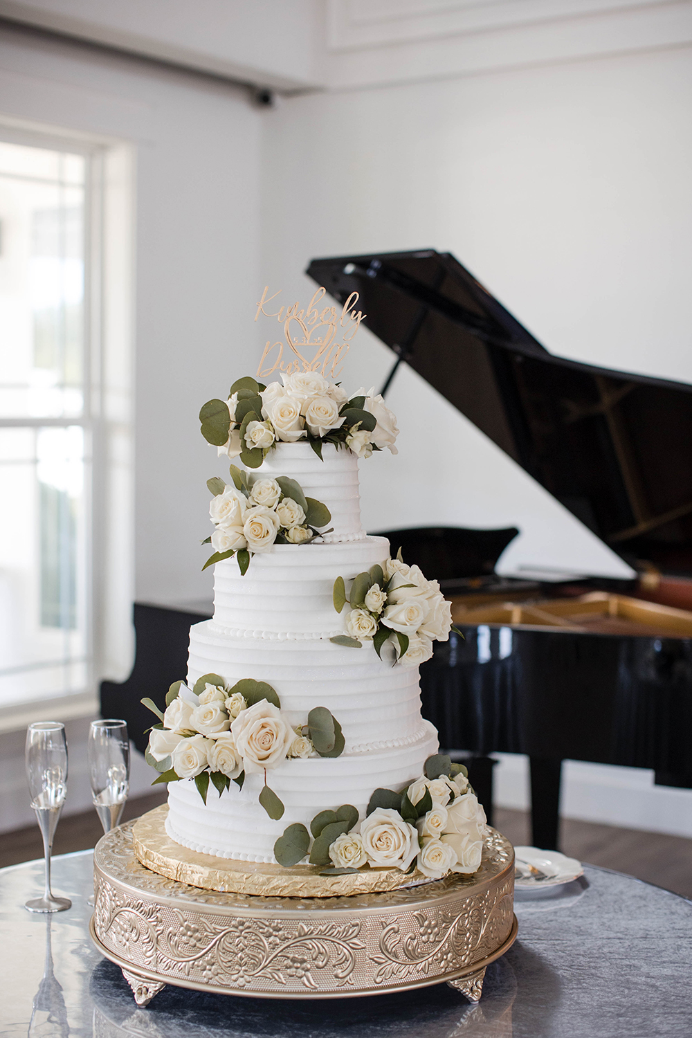 Three-tier white wedding cake decorated with white roses and greenery, displayed on an ornate stand with a piano in the background.