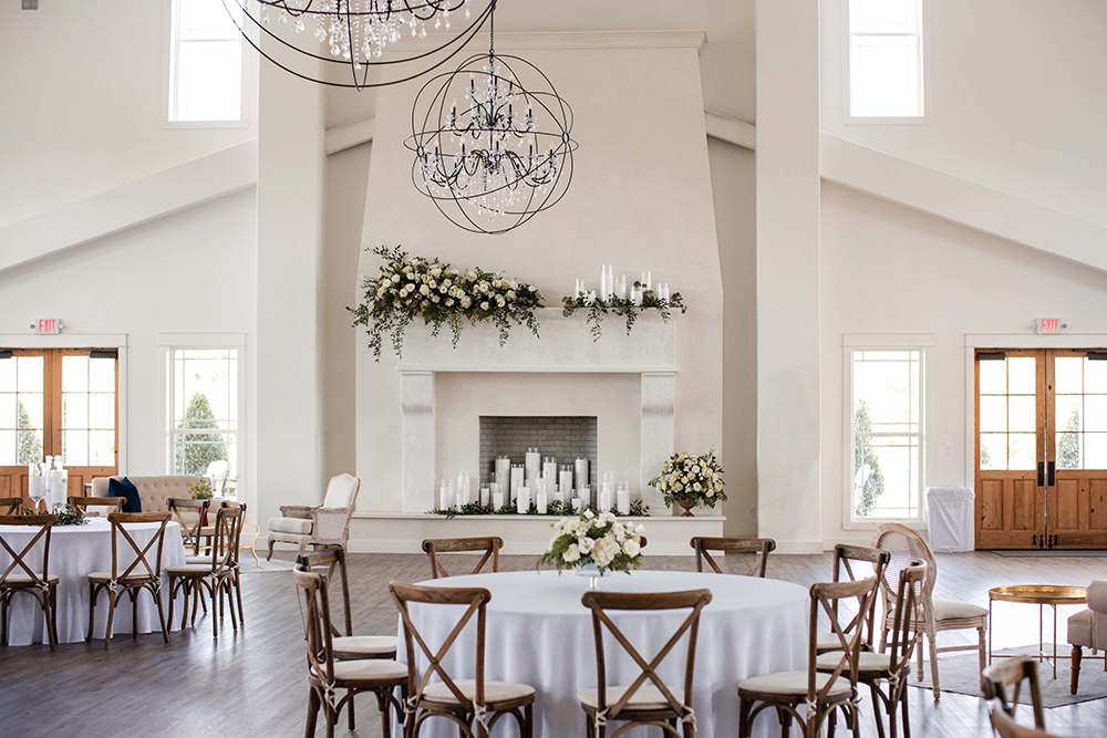 Elegant wedding reception space with round tables, wooden chairs, floral arrangements, and a large fireplace beneath hanging chandeliers.