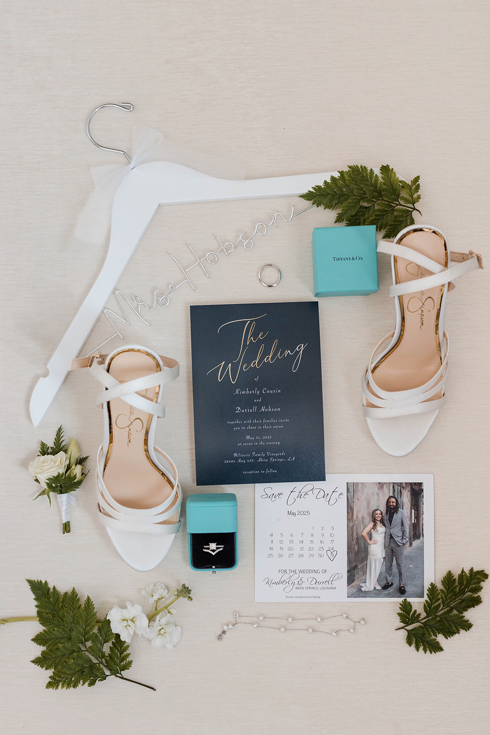 Flat lay of bride accessories including white heels, wedding invitation suite, save-the-date card, engagement ring, jewelry, and a personalized hanger on a light surface.