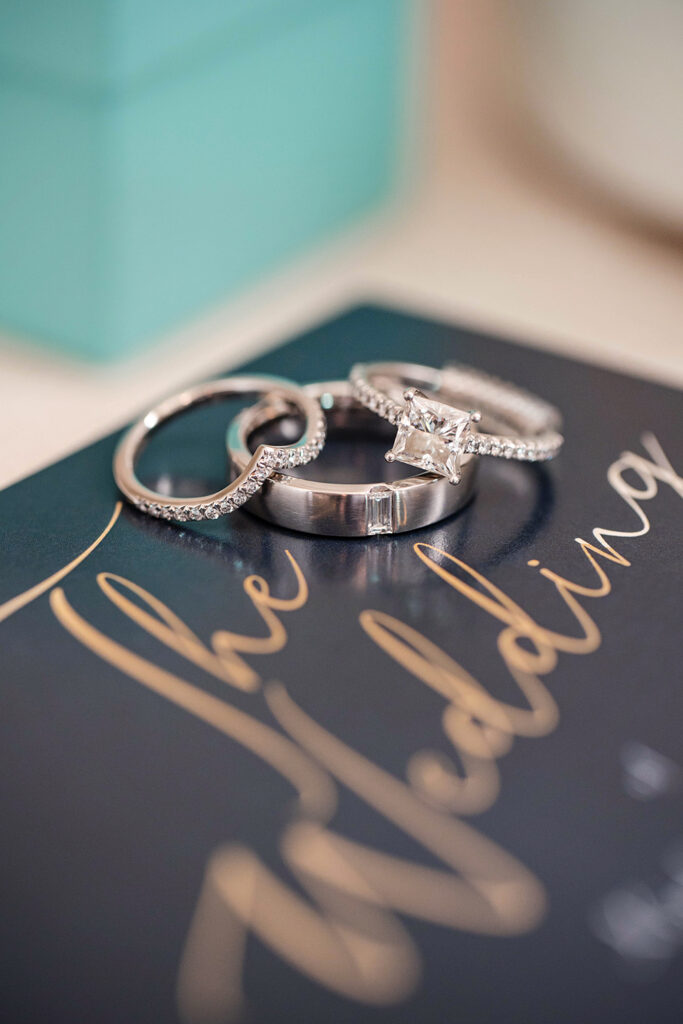 Close-up of wedding rings stacked on a wedding invitation, featuring a diamond engagement ring and matching bands.