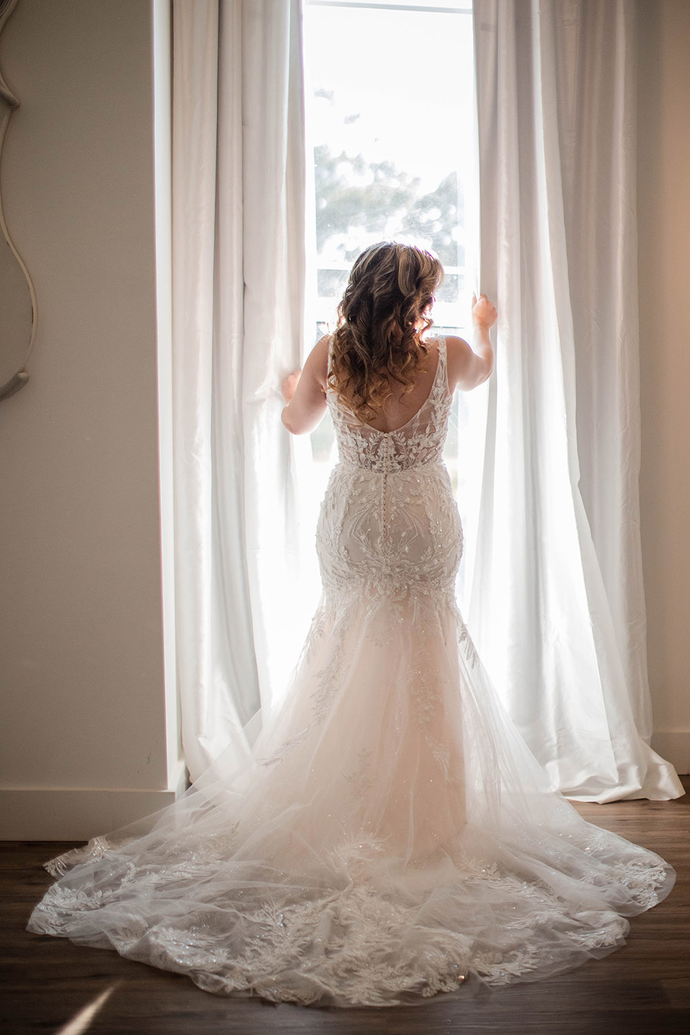 Bride in a lace wedding gown stands at a window, seen from behind, with sheer curtains and natural light filling the room.