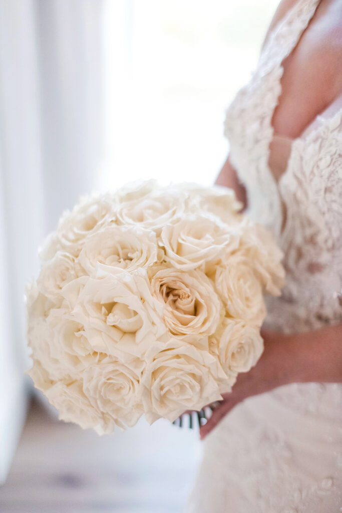 Close-up of a bride holding a round bouquet of white roses, with lace detail of her wedding gown visible.