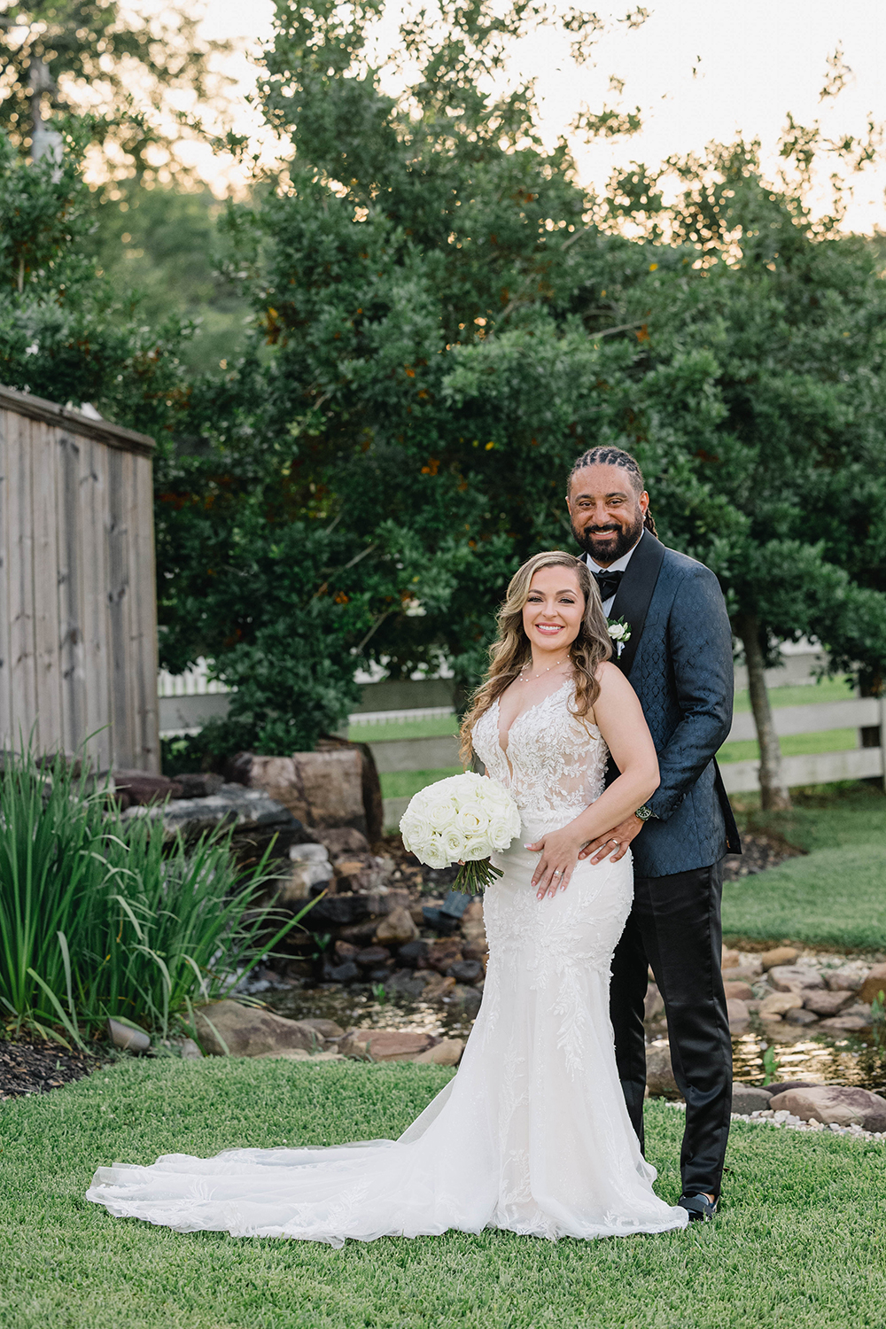 Bride in a lace wedding gown holding a white rose bouquet stands in front of the groom in a dark suit outdoors, both smiling at the camera.