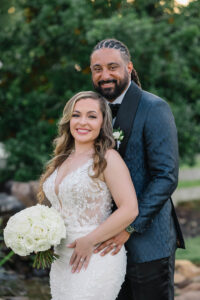 Bride in a lace wedding gown holding a white rose bouquet stands in front of the groom in a dark suit outdoors, both smiling at the camera.
