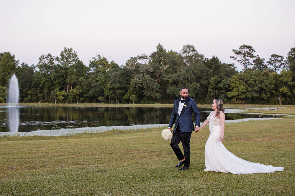 Bride and groom walk hand in hand across a grassy lawn beside a pond with a fountain, surrounded by trees.