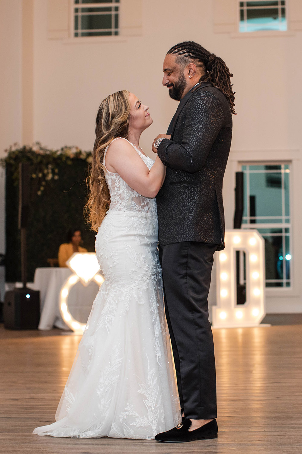 Bride and groom share their first dance on a wooden dance floor during the wedding reception, with illuminated initials in the background.