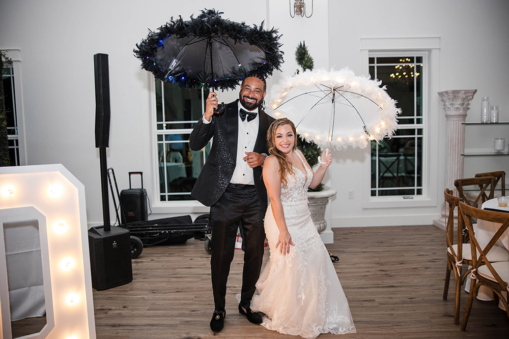 Bride and groom smile while holding decorative umbrellas during an indoor wedding reception, standing on a wooden dance floor.