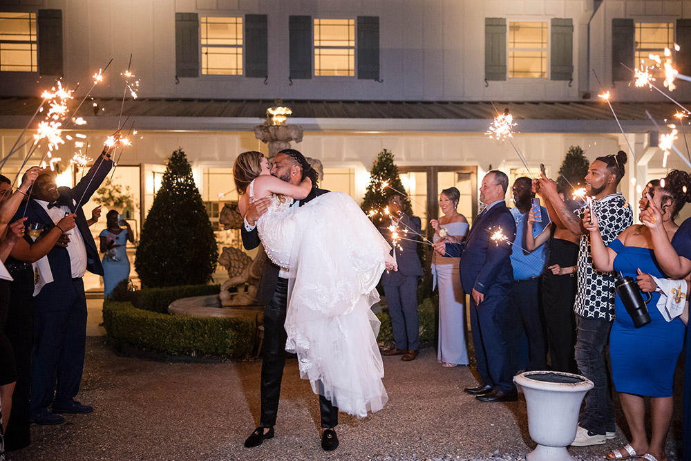 Groom lifts the bride during a nighttime sparkler exit, surrounded by wedding guests outside the reception venue.