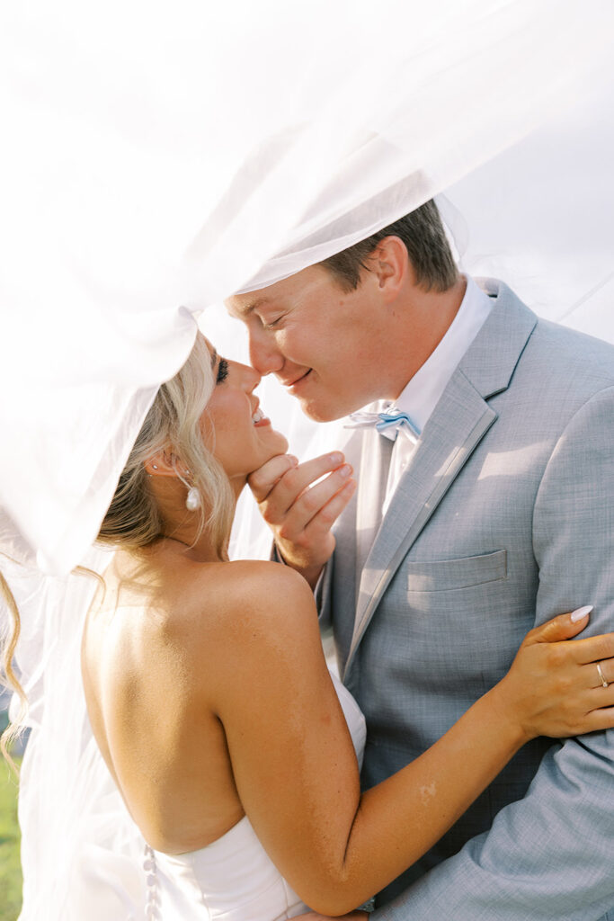 Close-up of a bride and groom embracing under the bride’s flowing veil, smiling as they lean in for a kiss during their wedding portraits.
