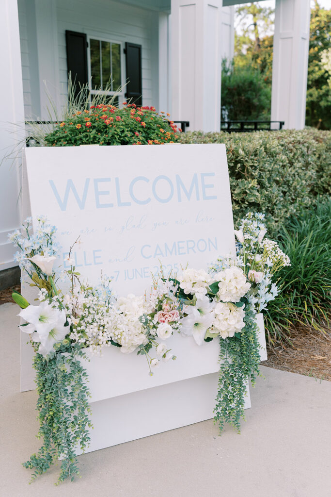 Wedding welcome sign decorated with white and pastel flowers and greenery, displayed outside a venue entrance, welcoming guests to Elle and Cameron’s June celebration.