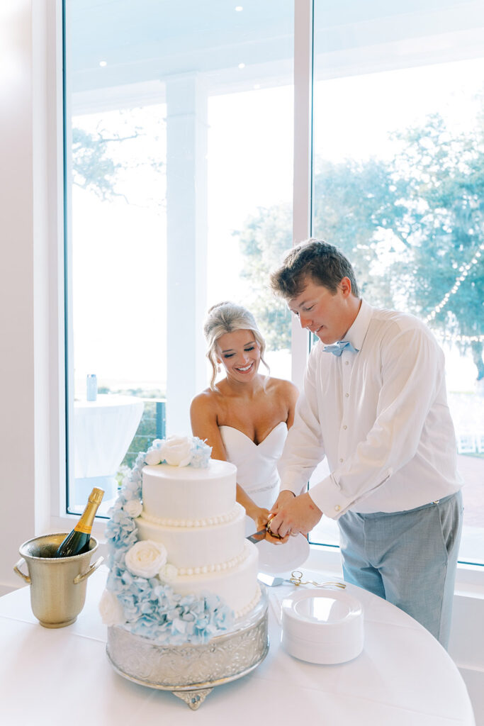 Bride and groom smiling as they cut their white tiered wedding cake decorated with blue and white flowers inside a bright reception venue with large windows.