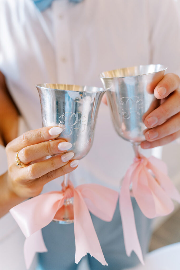 Close-up of a bride and groom holding two silver toasting goblets with monogram engraving and pink ribbon bows tied around the stems.