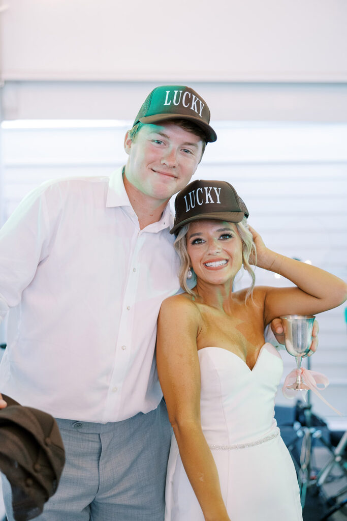 Bride and groom smiling at their wedding reception, both wearing matching hats that say “Lucky,” with the bride holding a silver goblet decorated with a pink ribbon.