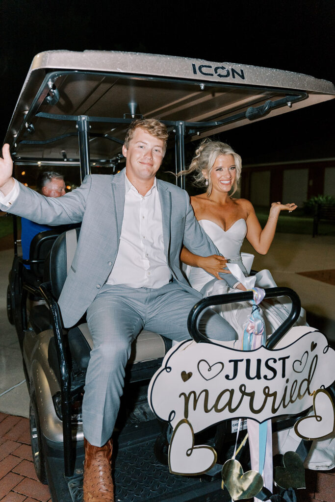 Bride and groom smiling and waving as they ride away in a golf cart decorated with a “Just Married” sign during their nighttime wedding exit.
