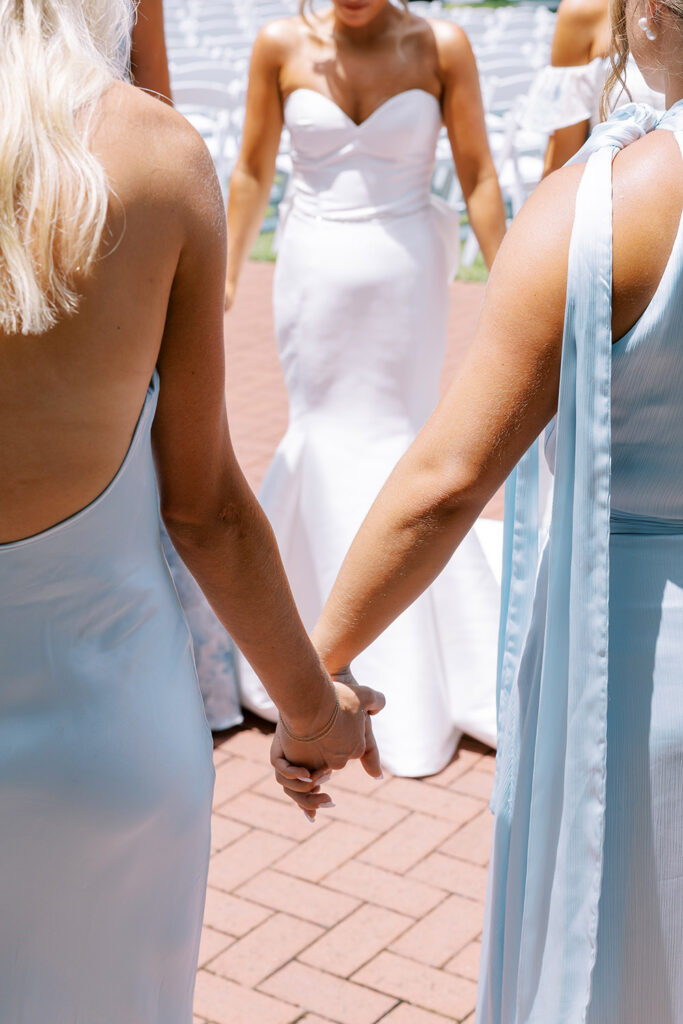 “Bride and bridesmaids holding hands in a prayer circle before the outdoor wedding ceremony, wearing white and light blue dresses.”