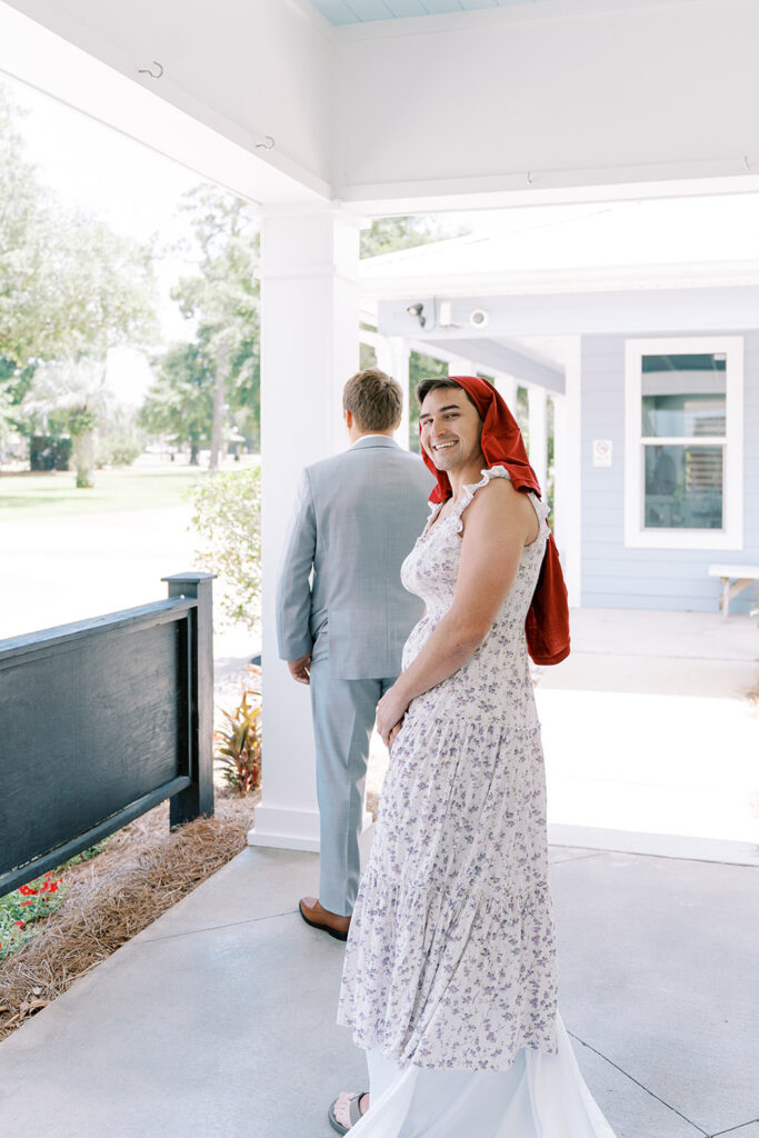 Groom standing with his back turned during a playful first-look moment as a man wearing a floral dress and red scarf smiles at the camera on a covered outdoor walkway.