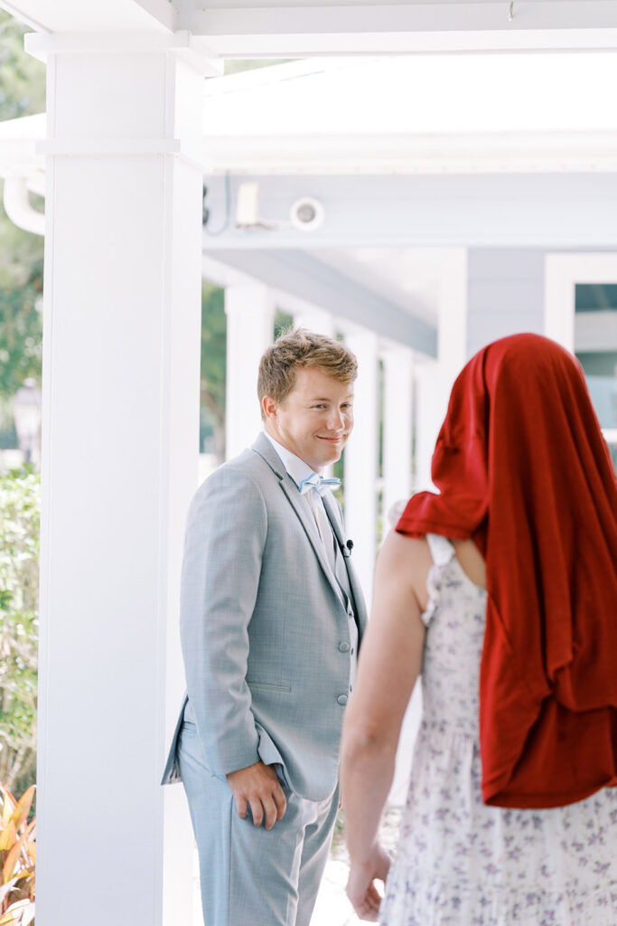 Groom in a light gray suit smiling as he turns to see a person playfully dressed in a floral dress with a red cloth over their head during a humorous first-look moment on a covered walkway.