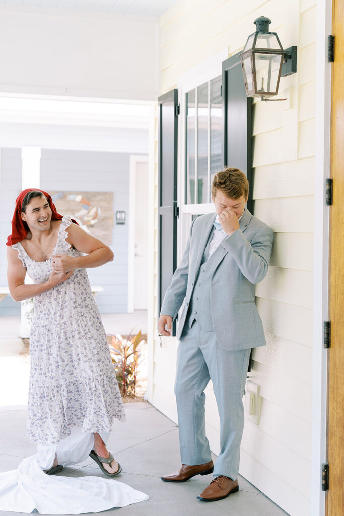 Groom in a light gray suit laughing and covering his face as a friend dressed humorously in a floral dress and red scarf pretends to be the bride during a playful first-look prank outside a light-colored building.