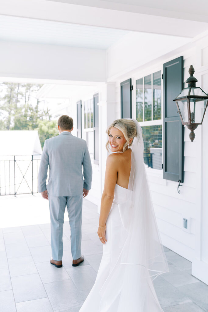 “Bride smiling at the camera before first look with groom in light gray suit on a white porch.”