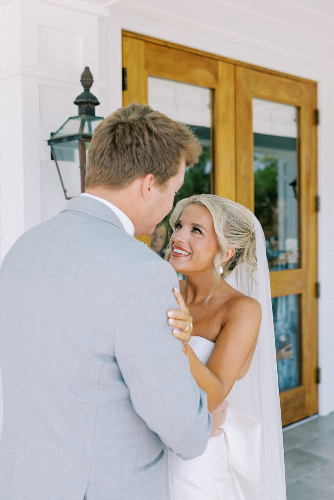 “Bride and groom share emotional first look on bright white porch, bride smiling in strapless wedding dress.”