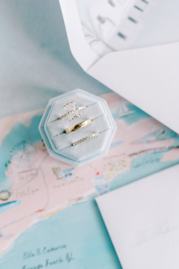 “Close-up of emerald-cut diamond engagement ring and wedding bands in a light blue velvet ring box, styled with coastal wedding invitation details.”